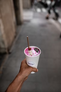 A hand holding a white cup of a fruit-based dessert or smoothie topped with granola, seeds, and coconut flakes. The dessert is a bright pink color, likely indicating ingredients such as berries or dragon fruit. A brown straw is placed in the cup. The background shows a blurred street, suggesting an outdoor urban setting.
