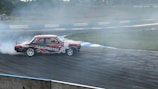 A vintage car with 'NANOPROTEC' branding is drifting on a race track, creating a plume of white smoke. The track has visible tire marks and is surrounded by blue and white barriers. The scene captures the dynamic motion and excitement of motorsport.