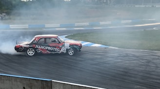 A vintage car with 'NANOPROTEC' branding is drifting on a race track, creating a plume of white smoke. The track has visible tire marks and is surrounded by blue and white barriers. The scene captures the dynamic motion and excitement of motorsport.