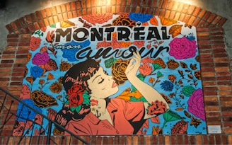 An artwork depicts a woman in profile with dark hair and a red rose tucked behind her ear. She holds a small object, possibly a cigarette, with painted nails. The background is filled with vibrant flowers in pink, orange, and blue hues. Above her, bold letters spell out 'Montreal mon amour.' The image is framed with brick architecture, suggesting an urban environment.