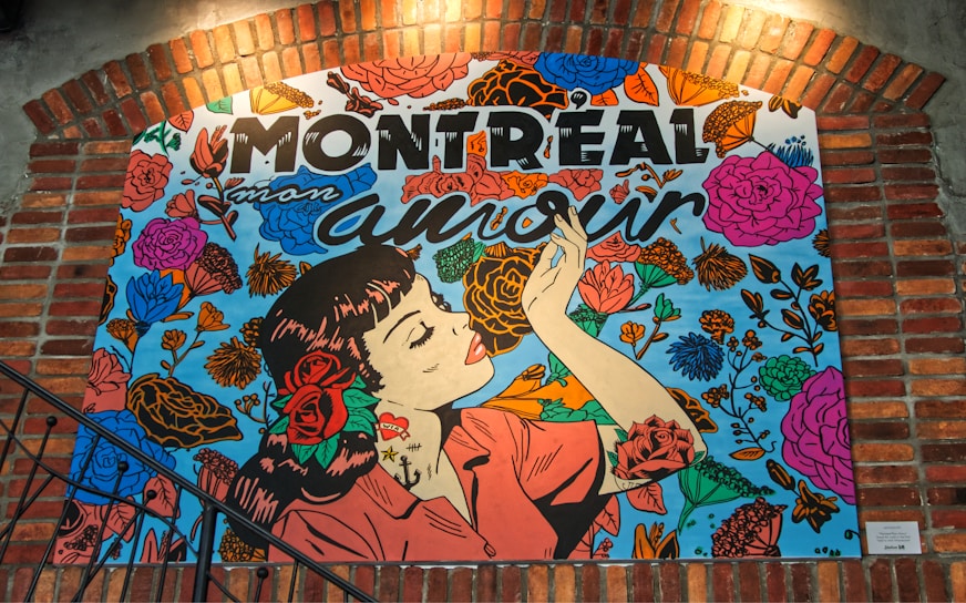 An artwork depicts a woman in profile with dark hair and a red rose tucked behind her ear. She holds a small object, possibly a cigarette, with painted nails. The background is filled with vibrant flowers in pink, orange, and blue hues. Above her, bold letters spell out 'Montreal mon amour.' The image is framed with brick architecture, suggesting an urban environment.