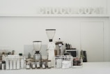 A minimalistic coffee counter with various coffee-making equipment prominently displayed. Two large coffee grinders are at the forefront, surrounded by glass jars filled with coffee beans and a selection of coffee packets stored in a wire basket. The backdrop features the words 'CHOCO COZY' in decorative letters. The overall setting is clean and organized, suggesting a focus on coffee preparation.