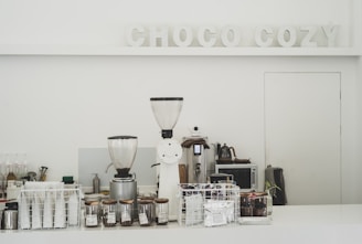 A minimalistic coffee counter with various coffee-making equipment prominently displayed. Two large coffee grinders are at the forefront, surrounded by glass jars filled with coffee beans and a selection of coffee packets stored in a wire basket. The backdrop features the words 'CHOCO COZY' in decorative letters. The overall setting is clean and organized, suggesting a focus on coffee preparation.