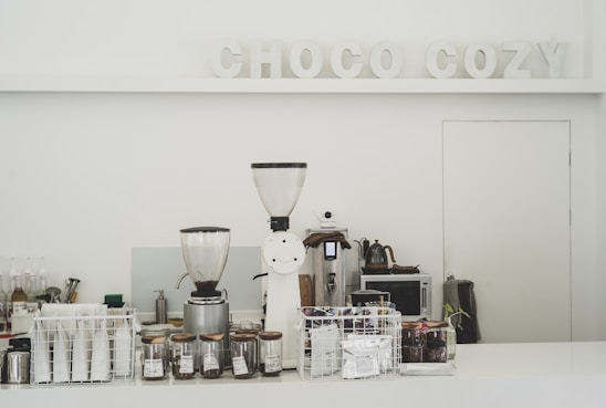 A minimalistic coffee counter with various coffee-making equipment prominently displayed. Two large coffee grinders are at the forefront, surrounded by glass jars filled with coffee beans and a selection of coffee packets stored in a wire basket. The backdrop features the words 'CHOCO COZY' in decorative letters. The overall setting is clean and organized, suggesting a focus on coffee preparation.