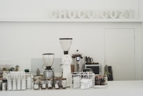 A minimalistic coffee counter with various coffee-making equipment prominently displayed. Two large coffee grinders are at the forefront, surrounded by glass jars filled with coffee beans and a selection of coffee packets stored in a wire basket. The backdrop features the words 'CHOCO COZY' in decorative letters. The overall setting is clean and organized, suggesting a focus on coffee preparation.