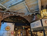 A cozy indoor marketplace with a rustic, industrial design featuring exposed brick walls and a high ceiling with visible pipes and beams. There are soft, warm lights hanging, and the entrance has a sign for a seafood buffet restaurant called Fisherman's Market Kobe. A display of stacked cans in vibrant colors is visible on one side, and fairy lights decorate the area.