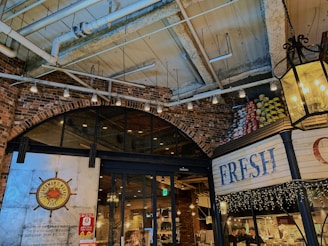 A cozy indoor marketplace with a rustic, industrial design featuring exposed brick walls and a high ceiling with visible pipes and beams. There are soft, warm lights hanging, and the entrance has a sign for a seafood buffet restaurant called Fisherman's Market Kobe. A display of stacked cans in vibrant colors is visible on one side, and fairy lights decorate the area.