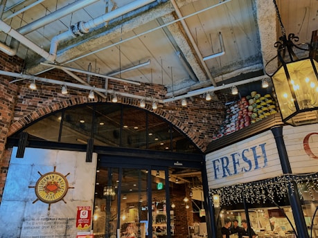 A cozy indoor marketplace with a rustic, industrial design featuring exposed brick walls and a high ceiling with visible pipes and beams. There are soft, warm lights hanging, and the entrance has a sign for a seafood buffet restaurant called Fisherman's Market Kobe. A display of stacked cans in vibrant colors is visible on one side, and fairy lights decorate the area.