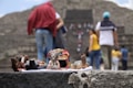 Several handmade clay figurines with vibrant paint are displayed on a stone surface. In the background, people are seen walking and engaging with what appears to be an ancient stone structure, suggesting a tourist or historical site.