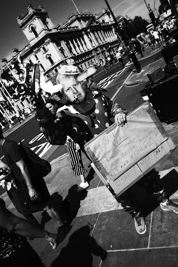 A person dressed in a clown costume holds a humorous sign with a list. The setting is an urban street with a historic building in the background and several pedestrians walking by. The image is captured in black and white.