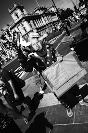 A person dressed in a clown costume holds a humorous sign with a list. The setting is an urban street with a historic building in the background and several pedestrians walking by. The image is captured in black and white.
