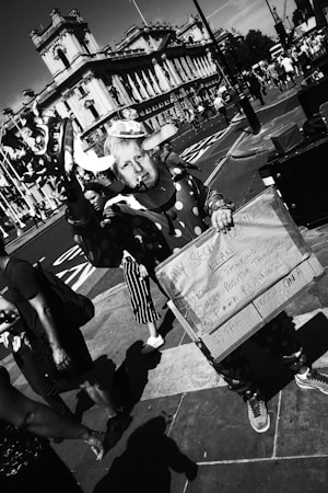 A person dressed in a clown costume holds a humorous sign with a list. The setting is an urban street with a historic building in the background and several pedestrians walking by. The image is captured in black and white.