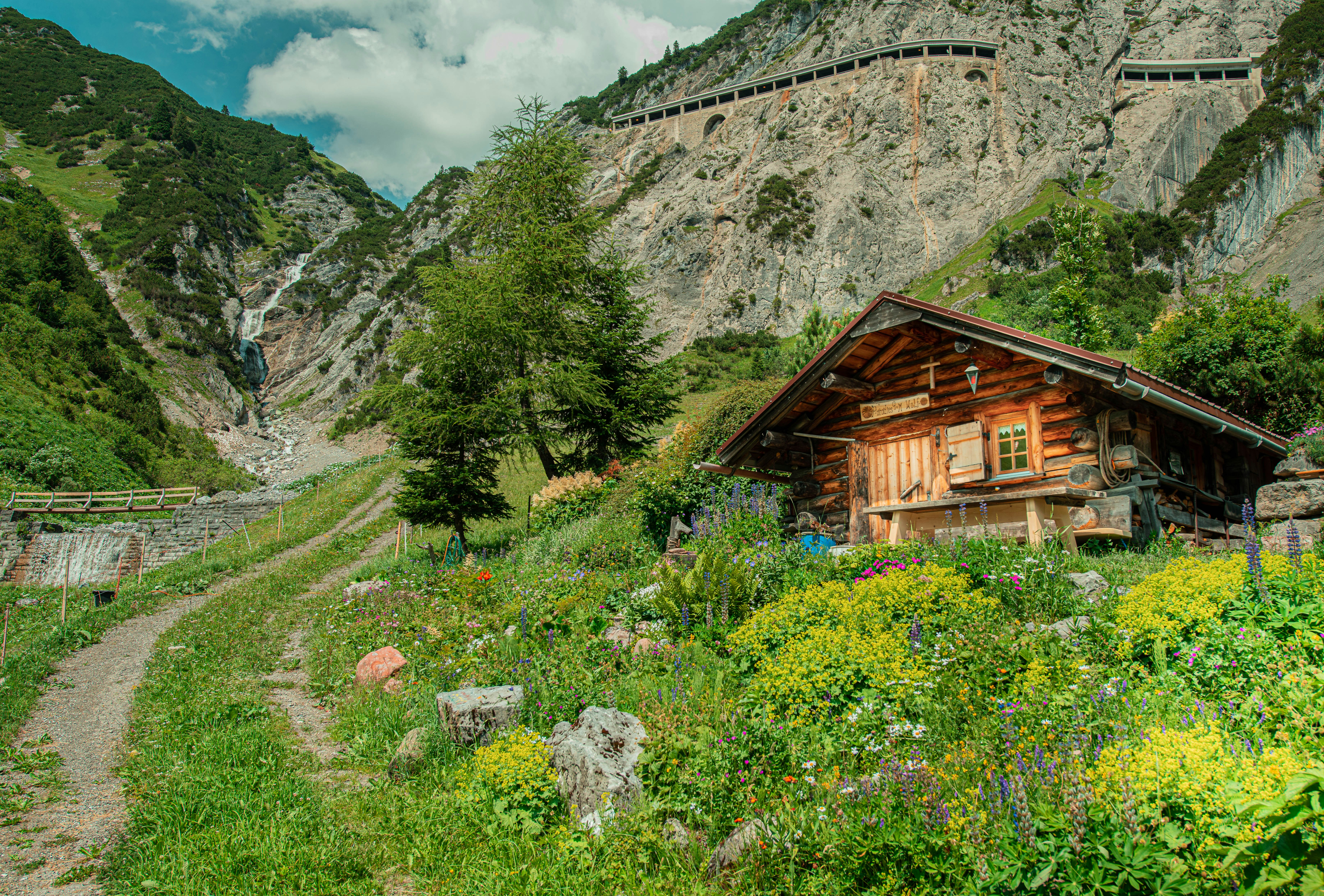brown wooden house near tree and mountain during daytime, A wonderful dreamy cottage just a few minutes outside of Sankt Anton, Austria. The incredible mountain road and the amazing waterfall in the background really thrilled me on this shot.