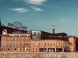 A historic red brick building with a vintage sign in Cyrillic script on its roof. The structure features multiple windows, a chimney, and modern glass additions. The sky is clear with some clouds.