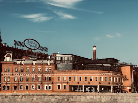 A historic red brick building with a vintage sign in Cyrillic script on its roof. The structure features multiple windows, a chimney, and modern glass additions. The sky is clear with some clouds.