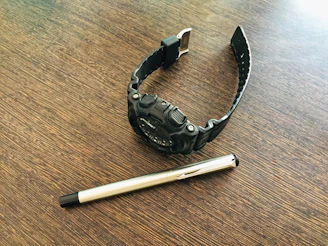 A close-up of a stylish men's watch resting on a wooden table beside a notebook and pen.