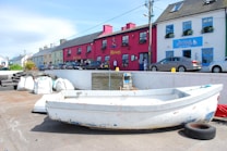 A small white boat is situated on a concrete dock, surrounded by items like tires and sandbags. In the background, a row of colorful, quaint houses painted in shades of red, blue, and beige line the street. The houses have signs suggesting they are pubs or cafes, with cars parked in front and a couple of people walking by.