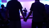 A group of people are gathered in a dimly lit room, holding hands. In the background, a person is speaking into a microphone on a stage with musical instruments around.