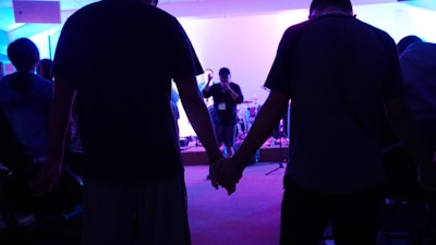 A group of people are gathered in a dimly lit room, holding hands. In the background, a person is speaking into a microphone on a stage with musical instruments around.