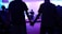 A group of people are gathered in a dimly lit room, holding hands. In the background, a person is speaking into a microphone on a stage with musical instruments around.