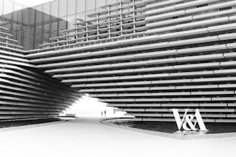 A modern architectural structure with overlapping horizontal layers creating a striking geometric pattern. The building features reflective glass panels and a pathway running underneath, leading to a bright opening in the distance. Two small figures walk along the path, emphasizing the scale of the structure. The letters 'V&A' are prominently displayed near the bottom right.