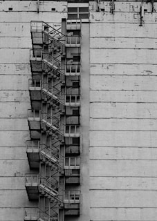A black and white image showing a tall building with an external zigzagging fire escape staircase. The staircase consists of metal stairs and platforms that protrude from the side of the building. The building has a worn, industrial appearance with visible signs of weathering and wear.