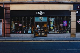 A storefront with black signage displays the logo for a shop named 'Beyond' with the number 88. The large glass windows reveal a glimpse inside, where various items and posters are visible. Strings of lights hang down, adding a festive touch. The sidewalk and street are in the foreground.