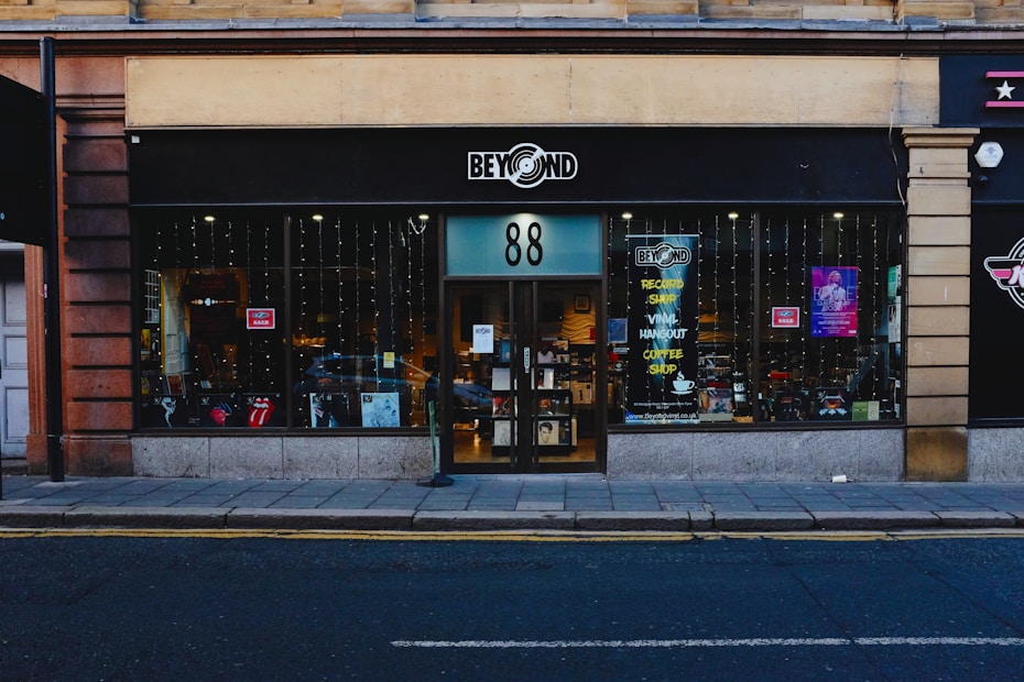 A storefront with black signage displays the logo for a shop named 'Beyond' with the number 88. The large glass windows reveal a glimpse inside, where various items and posters are visible. Strings of lights hang down, adding a festive touch. The sidewalk and street are in the foreground.