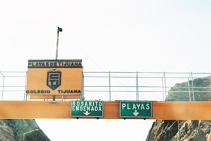 An overpass sign spans a road, prominently featuring two green directional signs below an orange information sign. The green signs read 'ROSARITO ENSENADA' and 'PLAYAS' with downward arrows. Above these, an orange sign with blue text reads 'PLAYAS DE TIJUANA COLEGIO TIJUANA,' alongside a logo. The structure is set against a bright, possibly sunny sky with rocky terrain visible to the sides.