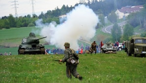 A World War II reenactment scene takes place in an open grassy field with several trees and rolling hills in the background. A tank is positioned to the left with soldiers dressed in period uniforms walking and engaging in simulated combat. Smoke billows from a simulated explosion near the center, indicating action. A military truck and armored vehicle are visible, along with a crowd of spectators watching the event from a distance.