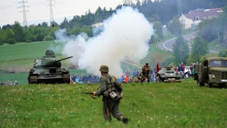 A World War II reenactment scene takes place in an open grassy field with several trees and rolling hills in the background. A tank is positioned to the left with soldiers dressed in period uniforms walking and engaging in simulated combat. Smoke billows from a simulated explosion near the center, indicating action. A military truck and armored vehicle are visible, along with a crowd of spectators watching the event from a distance.