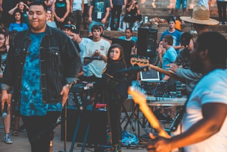 A lively outdoor concert or music event with people gathered around musicians performing. One person in the foreground appears focused while playing an instrument, and the crowd is engaged, some standing and others sitting.