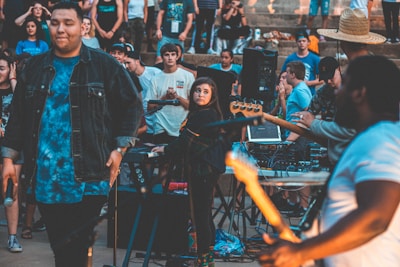A lively outdoor concert or music event with people gathered around musicians performing. One person in the foreground appears focused while playing an instrument, and the crowd is engaged, some standing and others sitting.