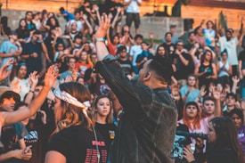 A large group of people, mostly young adults, are gathered outdoors with their hands raised. One person in the foreground, wearing a denim jacket, appears to be leading or addressing the crowd. There is a high level of engagement among the participants, some of whom are wearing bandanas or hats.