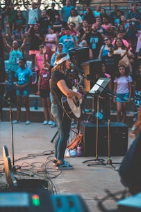 A musician performs onstage with a guitar and a microphone, surrounded by an enthusiastic crowd. The setting is outdoors, and people appear to be enjoying the live music event, with some raising their hands in excitement. The image captures a sense of community and engagement.