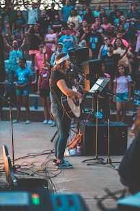 A musician performs onstage with a guitar and a microphone, surrounded by an enthusiastic crowd. The setting is outdoors, and people appear to be enjoying the live music event, with some raising their hands in excitement. The image captures a sense of community and engagement.