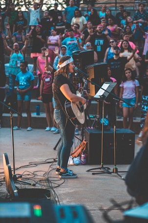 A musician performs onstage with a guitar and a microphone, surrounded by an enthusiastic crowd. The setting is outdoors, and people appear to be enjoying the live music event, with some raising their hands in excitement. The image captures a sense of community and engagement.