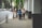 Two women walk down a city sidewalk next to an office building. One is pushing a stroller. A signboard advertises notary and mailing services. The street is lined with parked cars and trees.