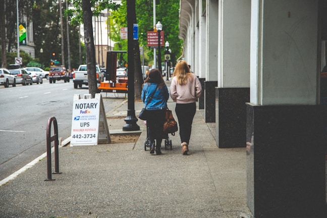 Two women walk down a city sidewalk next to an office building. One is pushing a stroller. A signboard advertises notary and mailing services. The street is lined with parked cars and trees.