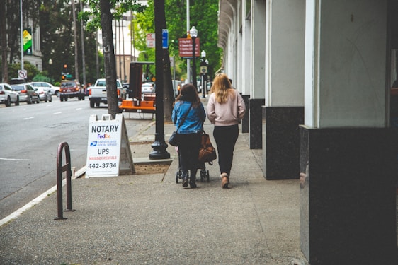 Two women walk down a city sidewalk next to an office building. One is pushing a stroller. A signboard advertises notary and mailing services. The street is lined with parked cars and trees.