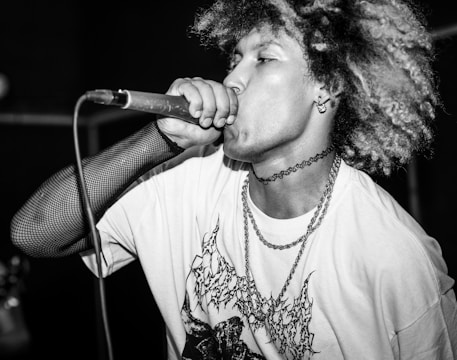 A performer passionately singing into a microphone, wearing a graphic t-shirt and fishnet-style arm sleeve. The individual has curly hair and is adorned with layered chains and a choker, conveying an energetic and expressive demeanor.