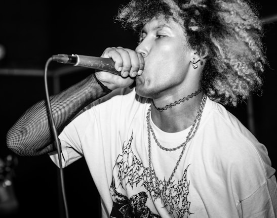 A performer passionately singing into a microphone, wearing a graphic t-shirt and fishnet-style arm sleeve. The individual has curly hair and is adorned with layered chains and a choker, conveying an energetic and expressive demeanor.