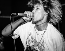 A performer passionately singing into a microphone, wearing a graphic t-shirt and fishnet-style arm sleeve. The individual has curly hair and is adorned with layered chains and a choker, conveying an energetic and expressive demeanor.