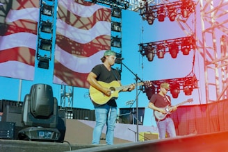 Musicians perform on an outdoor stage with large screens displaying a patterned backdrop. The setup includes microphones, guitars, and stage lights. The sky is clear, contributing to a vibrant atmosphere.