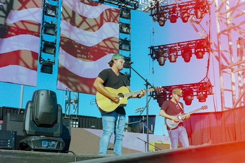 Musicians perform on an outdoor stage with large screens displaying a patterned backdrop. The setup includes microphones, guitars, and stage lights. The sky is clear, contributing to a vibrant atmosphere.