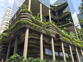 An urban architectural structure features multiple levels clad with lush green vegetation, interspersed with sleek, modern glass and metal elements. The building utilizes a blend of organic and contemporary design, highlighting vertical gardens and tiered plant arrangements.