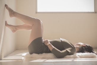 A person is lying on a soft surface inside a bright, airy room, with their legs raised against a wall and hands resting on their stomach. The room is filled with natural light coming from a window.
