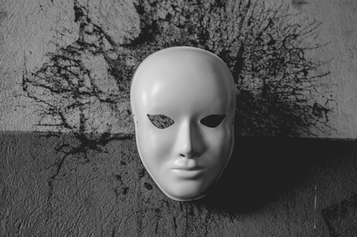 A white, featureless mask is prominently displayed against a textured wall with a dark, abstract splash pattern behind it. The mask's eyes are hollow, lending it a mysterious and slightly eerie aura. The entire image is in grayscale, enhancing the stark contrast between the mask and the background.