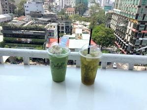 Two plastic cups with dome lids and straws, filled with green and yellow drinks, sit on a white ledge overlooking a cityscape. The background features urban buildings, lush trees, and a branded sign reading 'Saigon Garden.' The scene is captured from a high vantage point, showcasing the bustling city environment.
