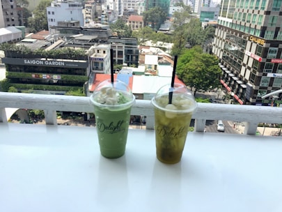 Two plastic cups with dome lids and straws, filled with green and yellow drinks, sit on a white ledge overlooking a cityscape. The background features urban buildings, lush trees, and a branded sign reading 'Saigon Garden.' The scene is captured from a high vantage point, showcasing the bustling city environment.